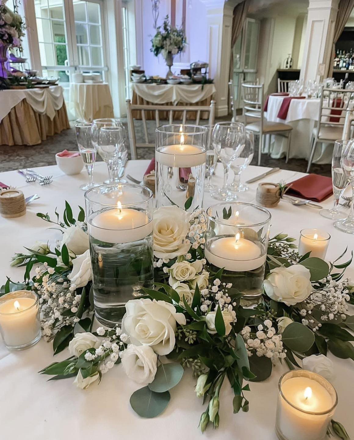 Clear glass cylinder vases with floating candles surrounded by white roses and greenery on a table.