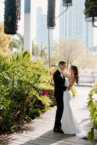 Outdoor portrait of a bride and groom embracing in a lush garden setting with Miami's skyline in the background, ca