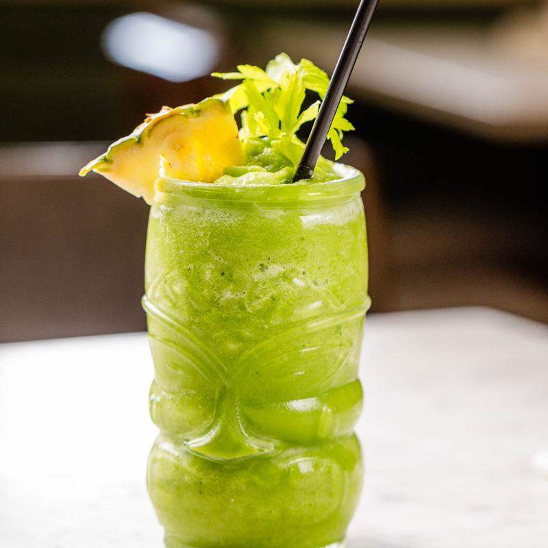 A vibrant green drink in a tiki glass, garnished with pineapple wedge and celery leaves.