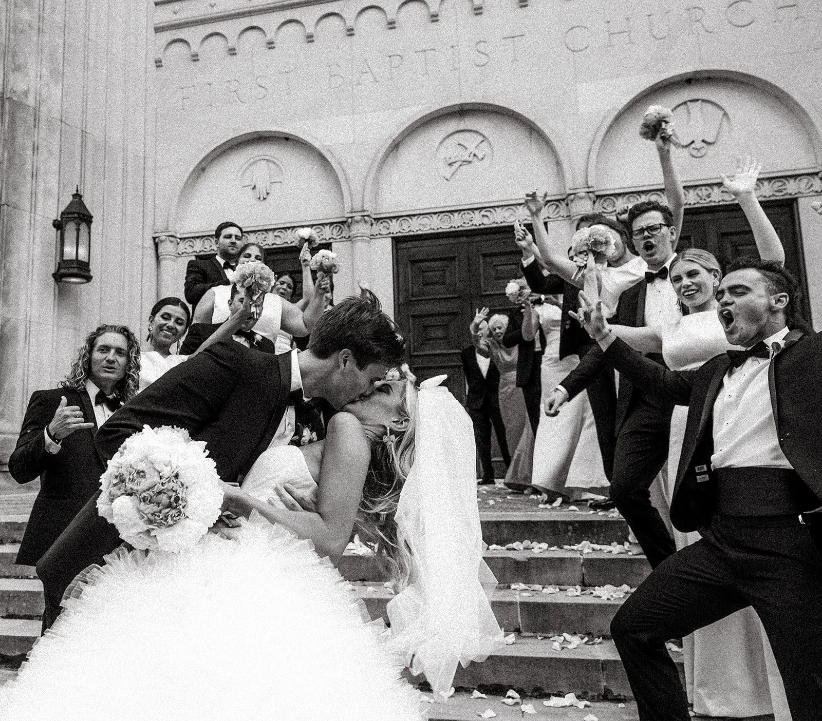 A wedding celebration with a bride and groom kissing on church steps surrounded by friends and family. The guests are ch
