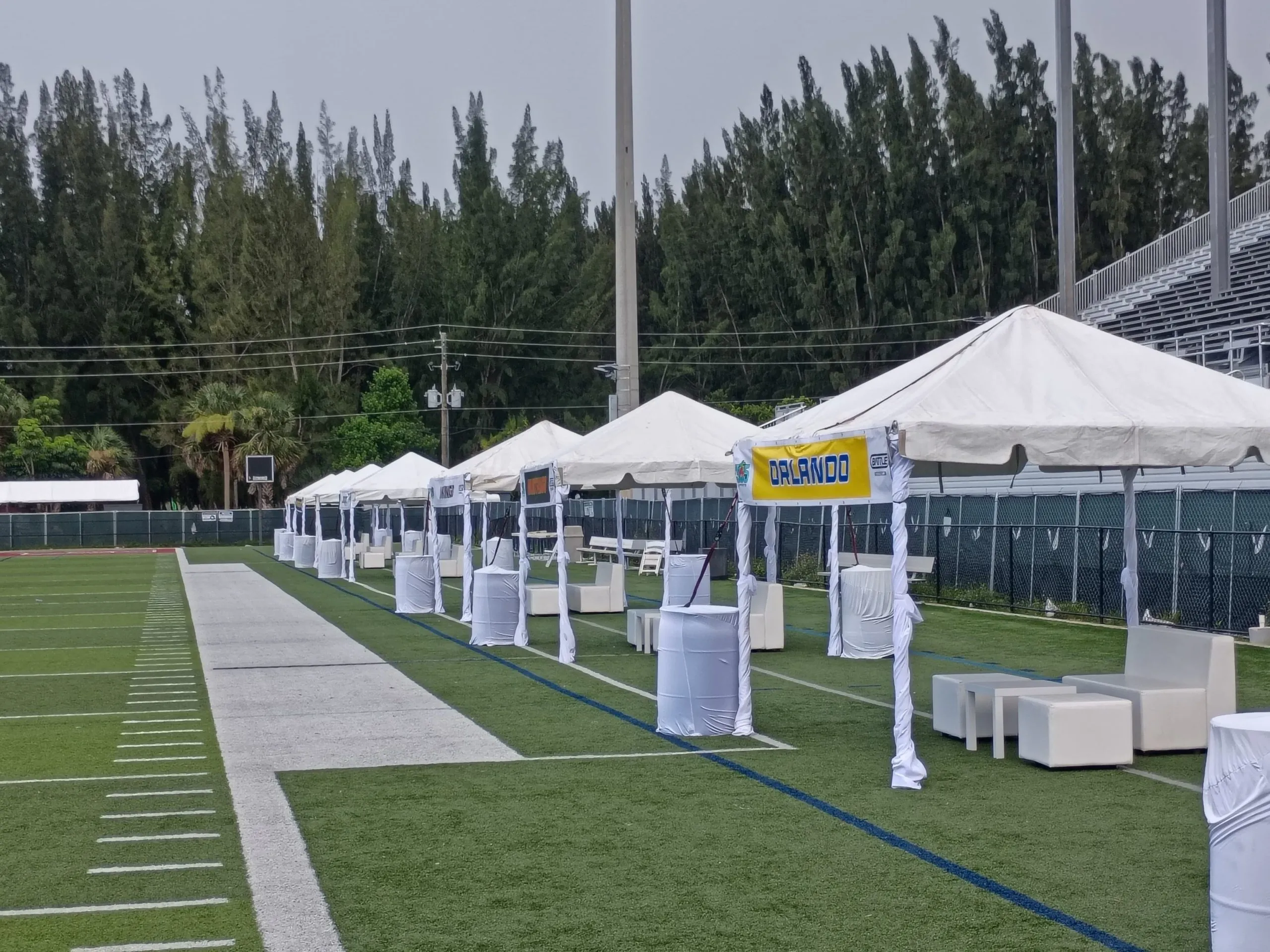 A row of white 10 by 10 event tents with lounge seating and cocktail tables beside a football field