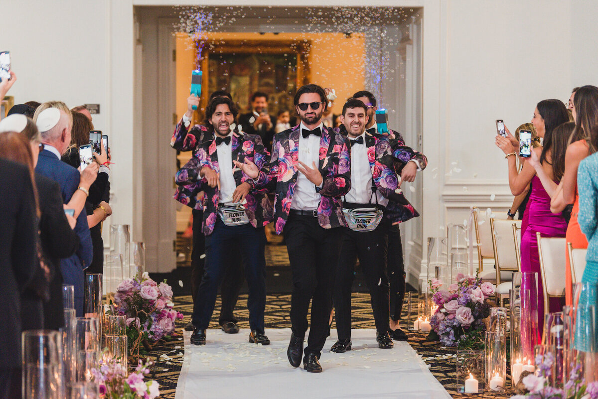Groomsmen dancing down the reception aisle during a lively wedding entrance surrounded by floral arrangements and cheeri