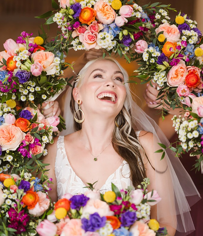 Bride laughing surrounded by colorful floral wreaths with vibrant roses and wildflowers.