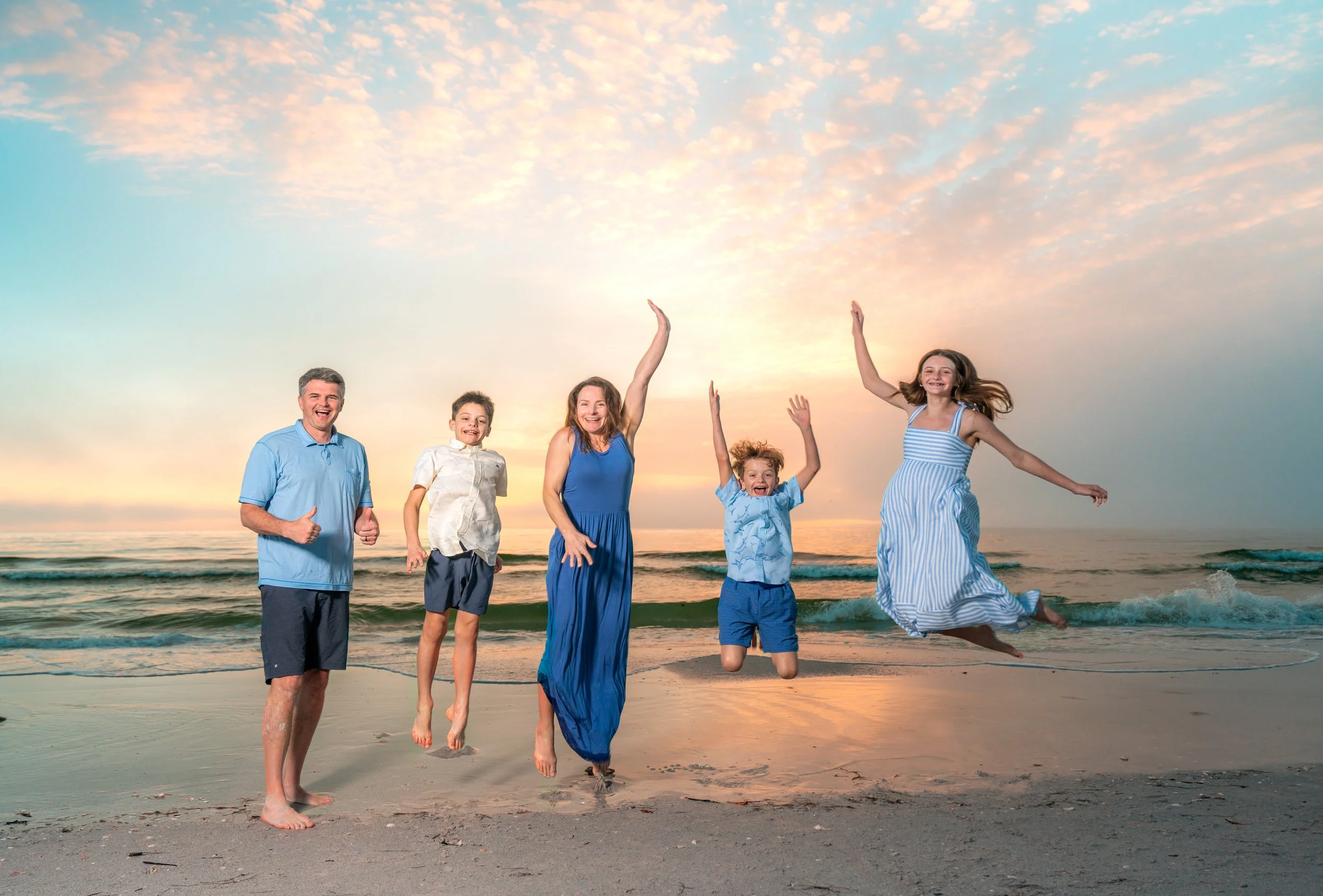 Family of five enjoying a sunny day at the beach, smiling and playing near the water with colorful sky in the background