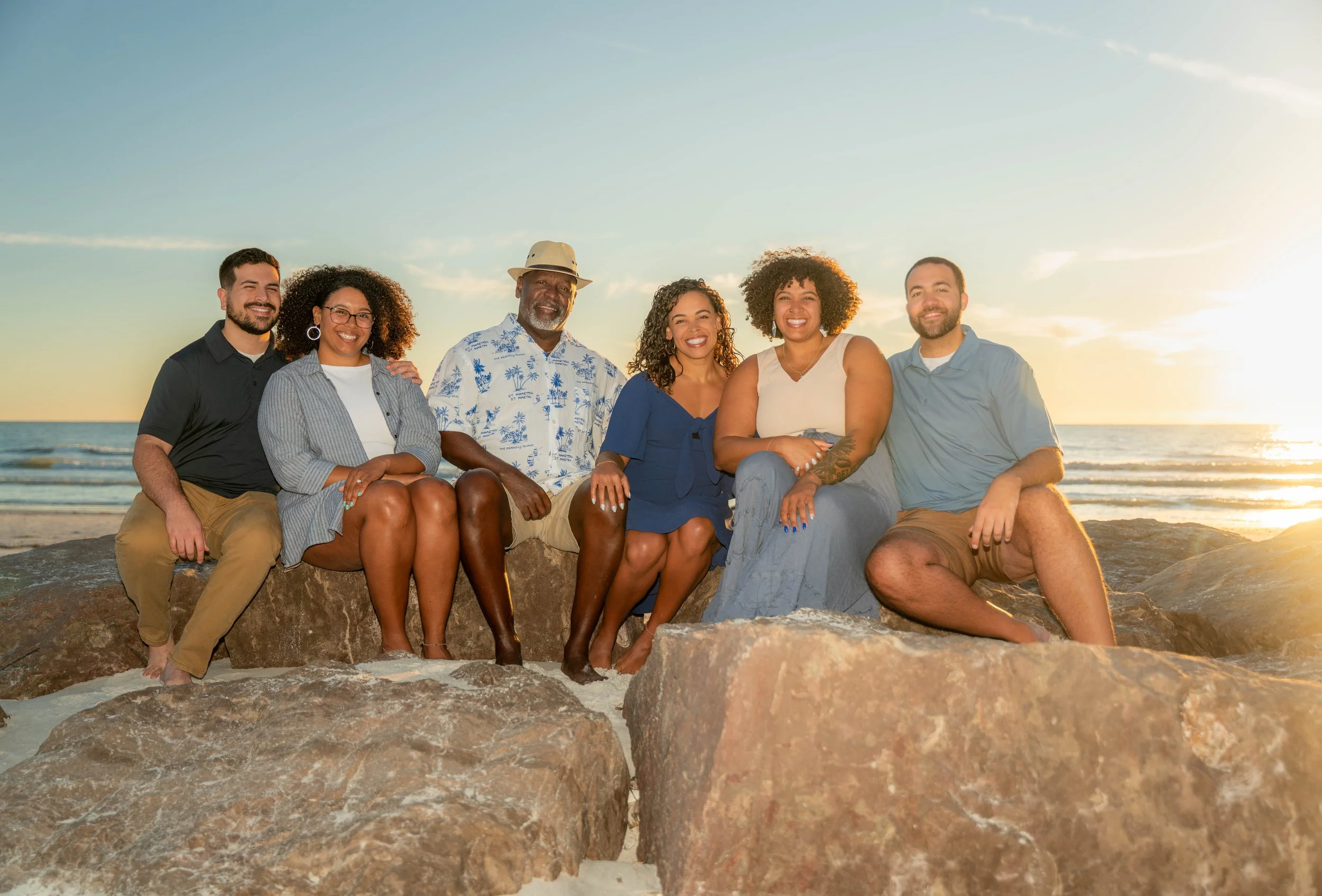 Group of six diverse friends sitting on rocks at the beach during sunset, smiling and enjoying the moment.