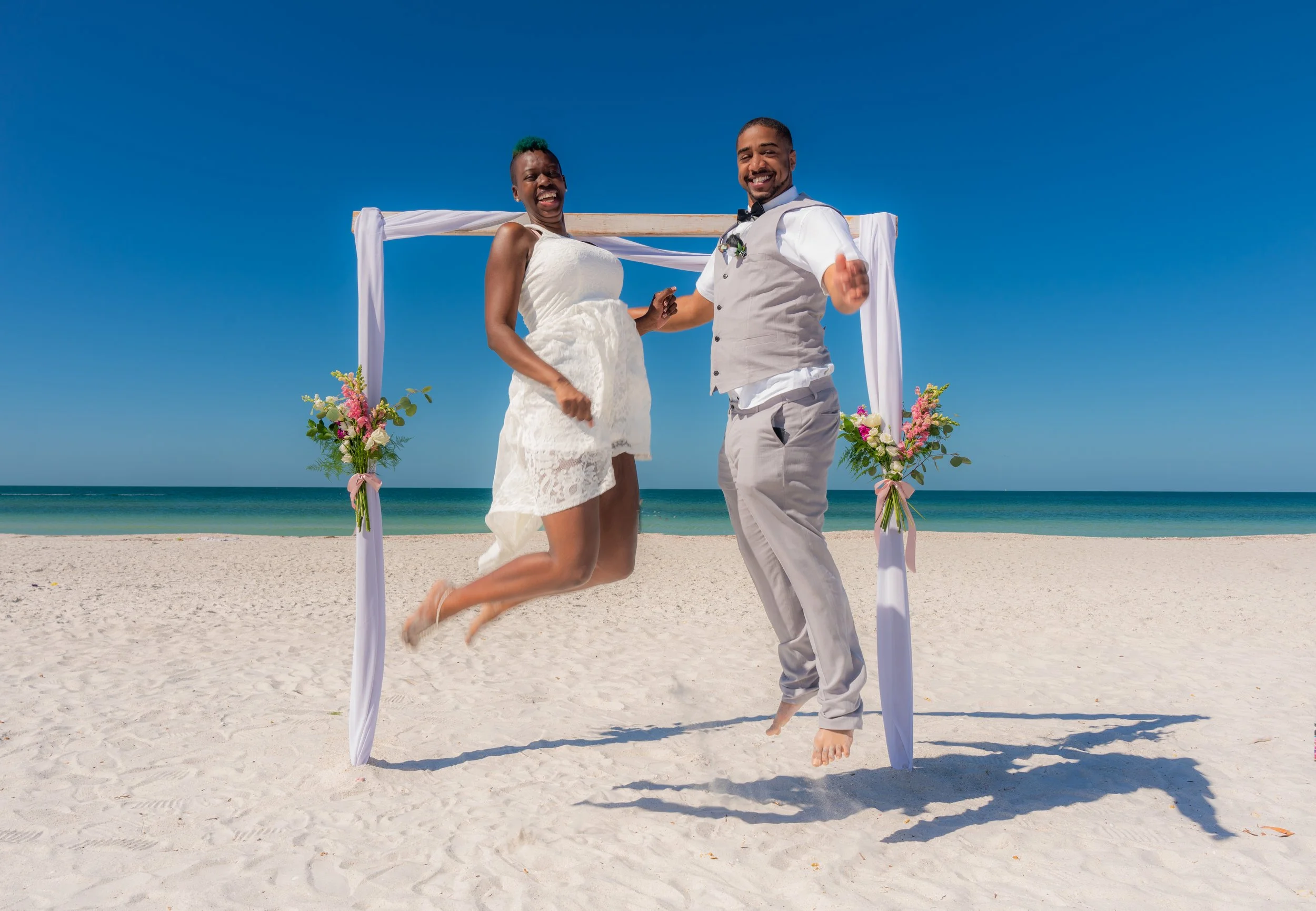 A newlywed couple at the beach, holding hands under an arch decorated with flowers, smiling and dancing on the sand with