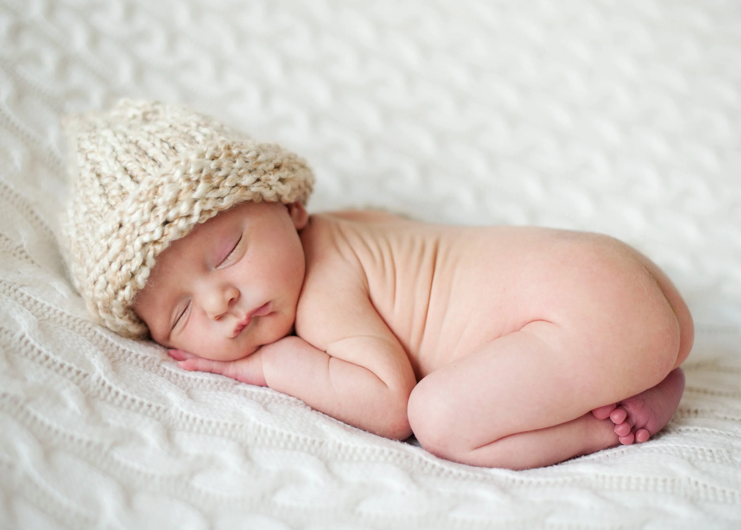 Sleeping newborn baby lying on a soft white blanket, wearing a knitted beige hat.