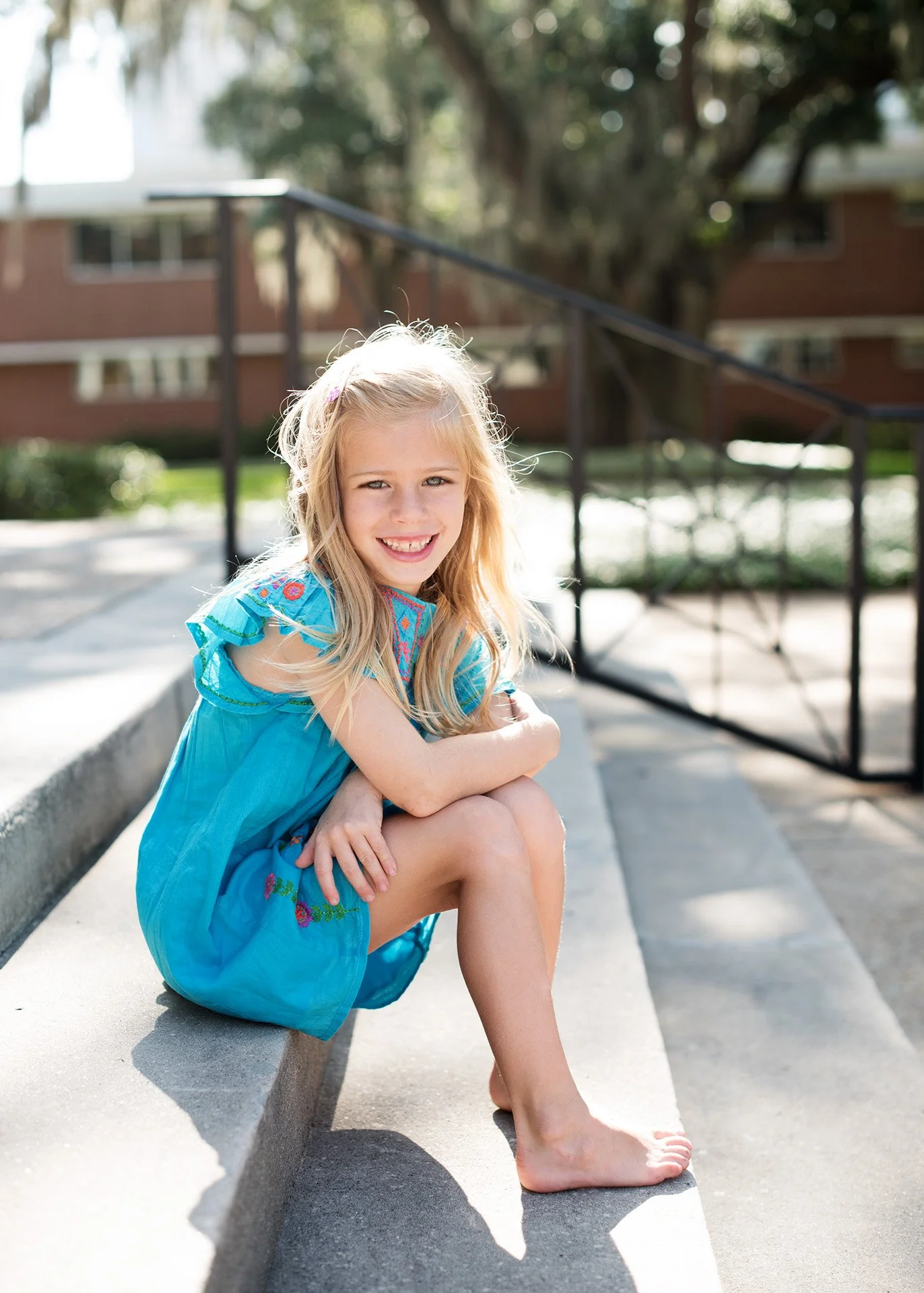 Young girl with long blond hair, sitting on concrete steps outdoors, wearing a blue dress, smiling, with a school buildi