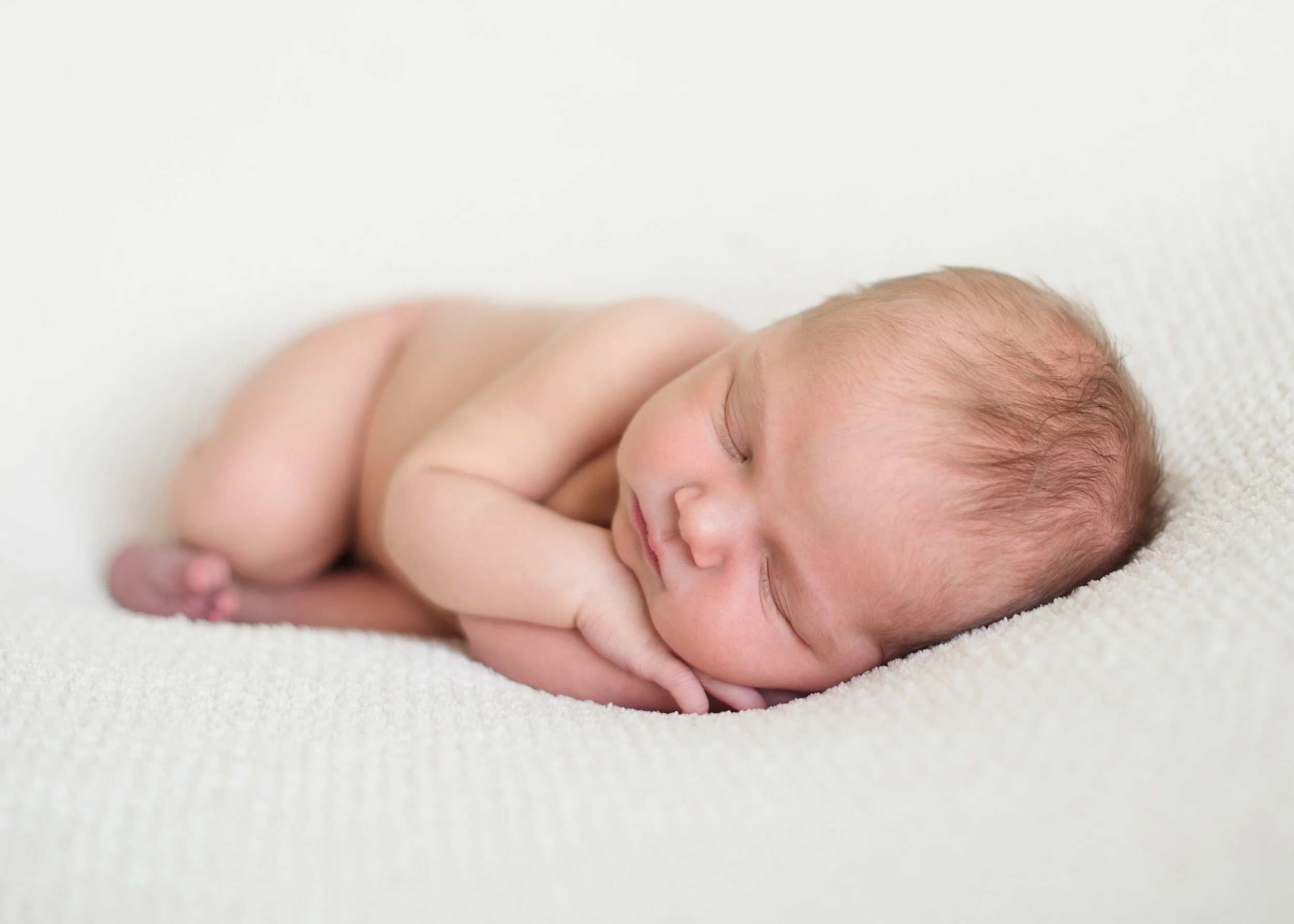 A sleeping newborn baby lying face down on a soft, cream-colored blanket with arms curled under its chin newborn session