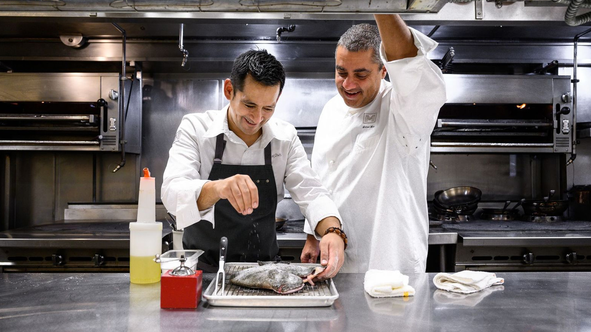 Two chefs stand in a modern kitchen. One chef, smiling, sprinkles seasoning on a whole fish on a metal tray, while the o