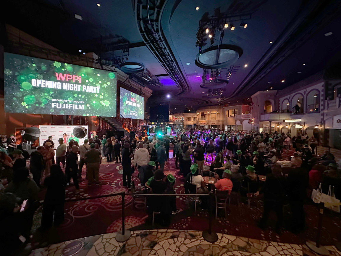 A large crowd at the WPPI opening night party inside a spacious venue with stage lights and a big screen.