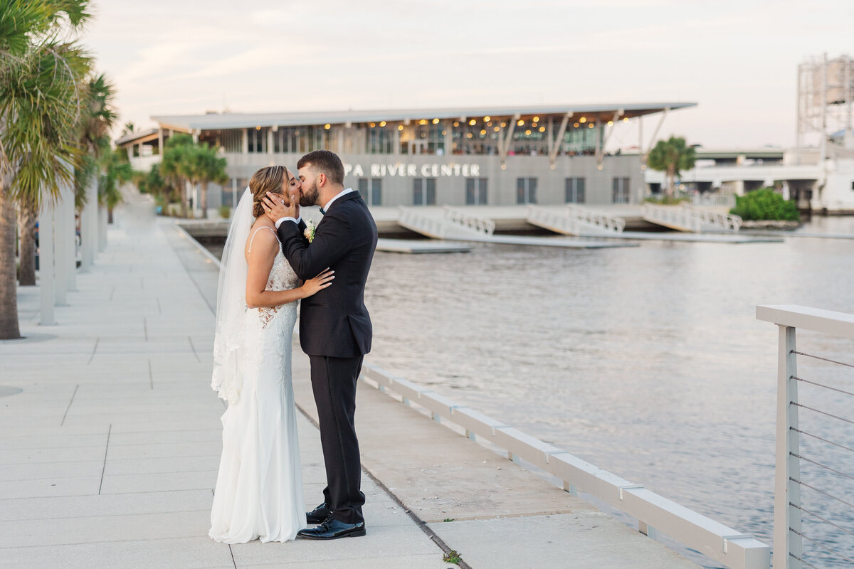 Bride and groom kiss on the riverfront walkway with Tampa River Center behind them on the Hillsborough River in Tampa