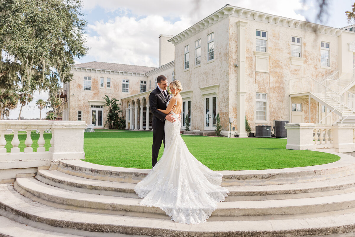 Bride and groom embrace on the curved stone steps at Bella Cosa in Lake Wales, Florida, her lace train spread down the s