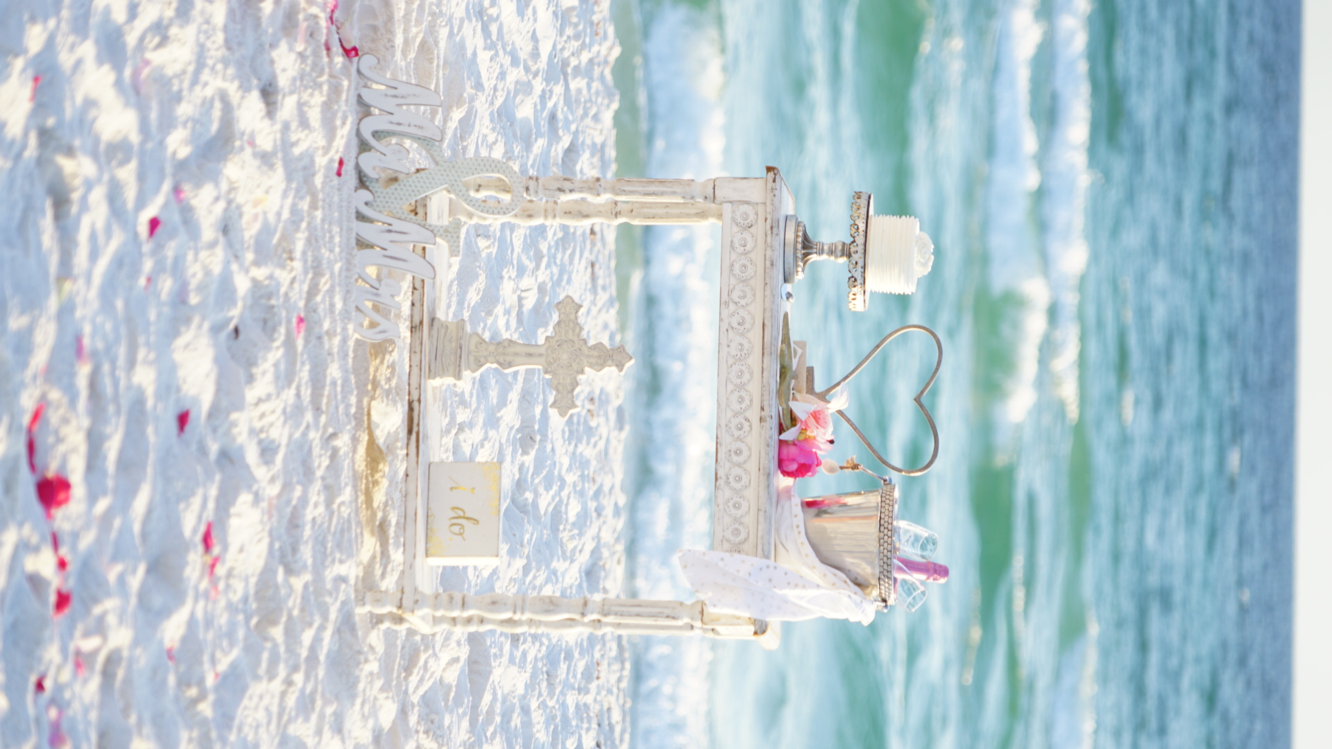 Luxury beach wedding setup with cake, champagne, heart decoration, cross, I Do sign, and Mr and Mrs sign on the white sa