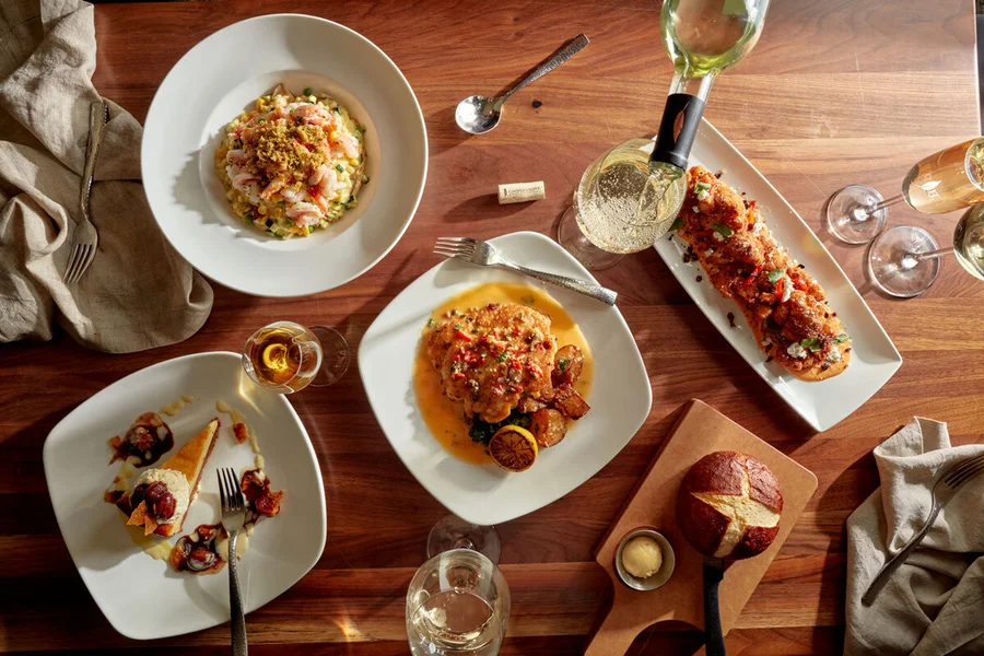Overhead view of a wooden table set with plated gourmet dishes, bread, wine glasses, and silverware, with warm sunlight