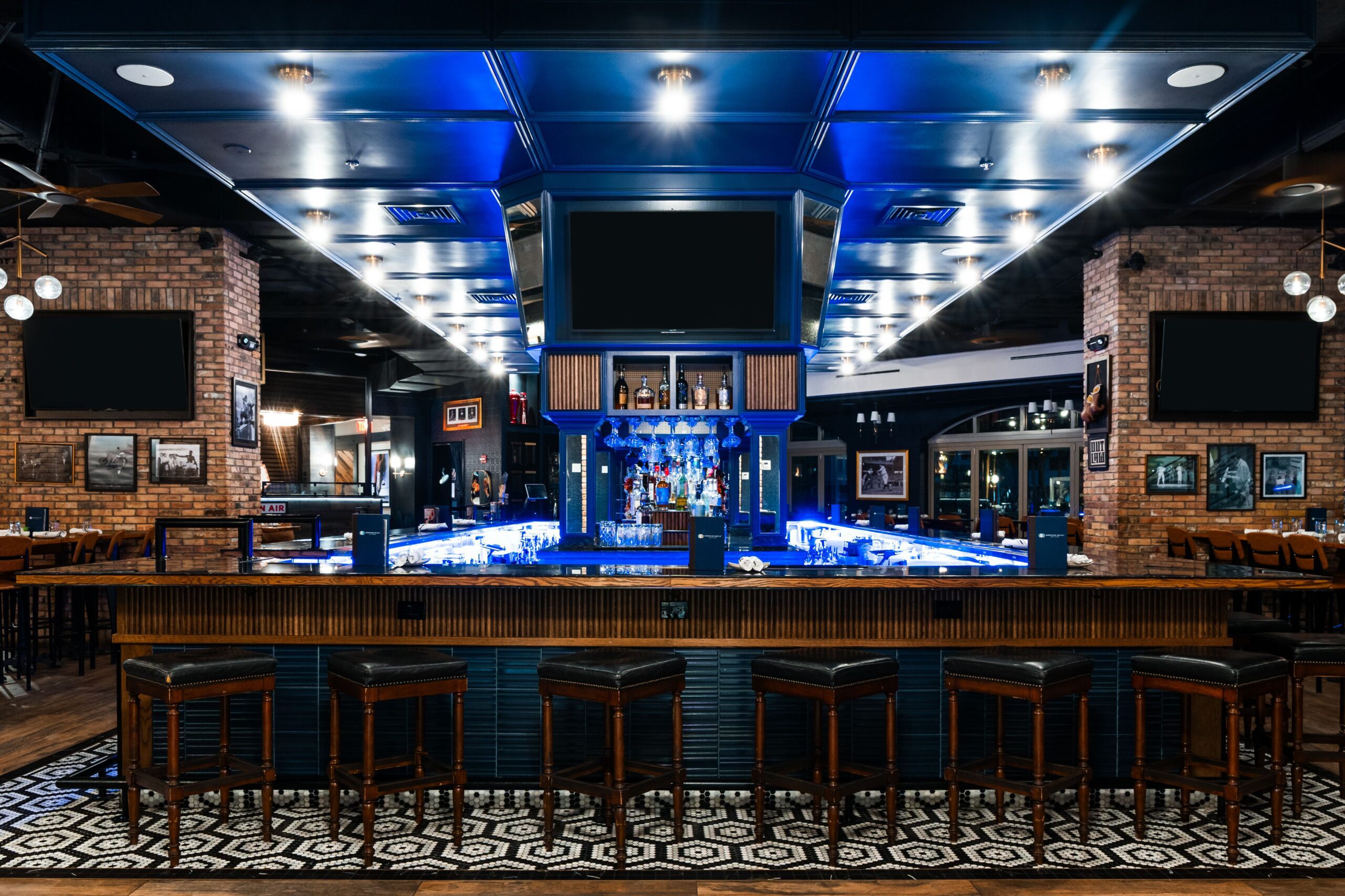 A modern Dining AmSo bar interior features a central bar counter surrounded by black barstools, illuminated shelves with