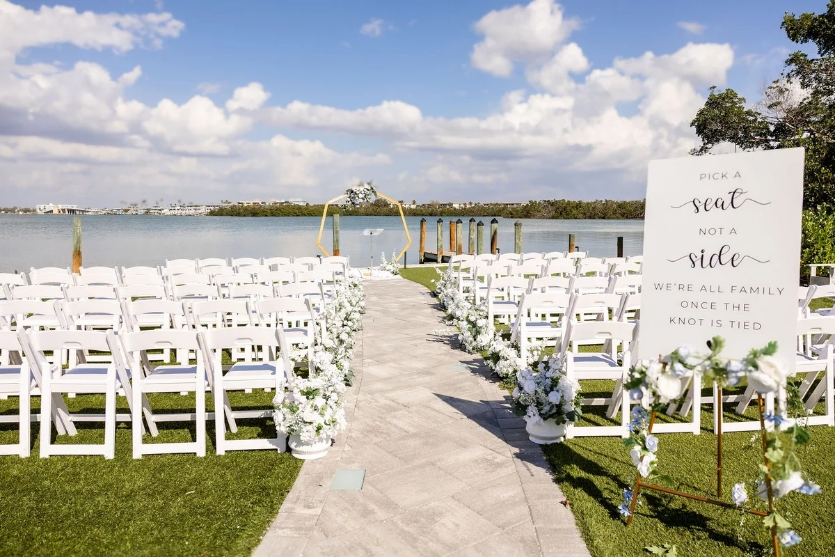 Rows of white chairs decorated with flowers are arranged facing a floral arch by Lemon Bay for an outdoor wedding in Eng