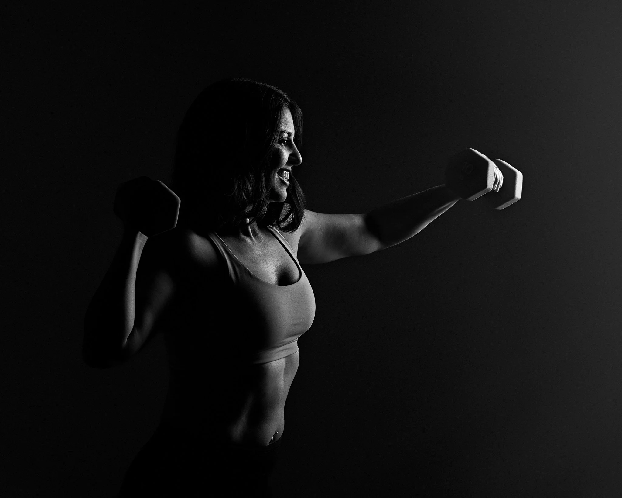 A woman in a workout pose, holding dumbbells in a dark setting, highlighting her muscular silhouette.