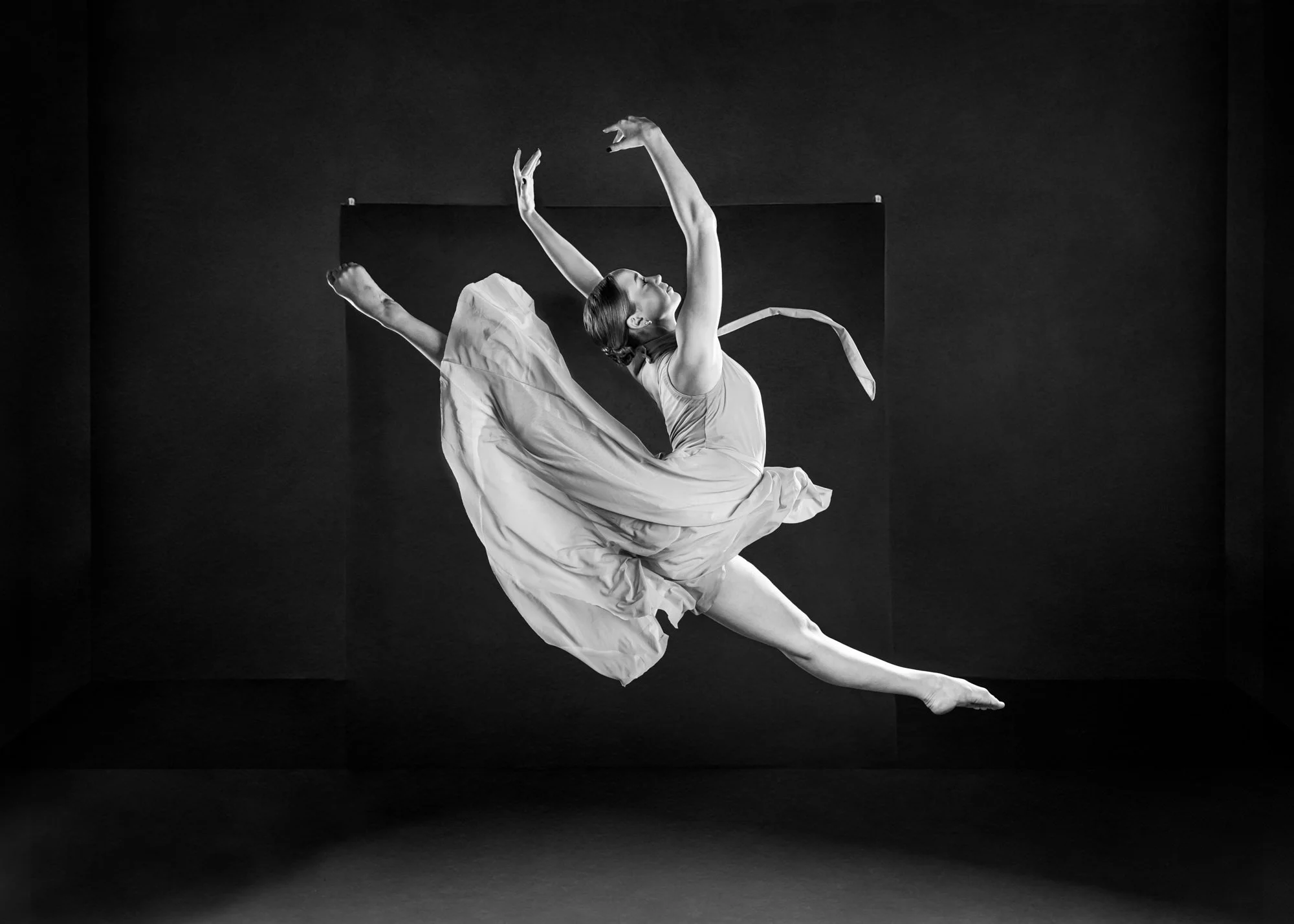 Black and white photo of a female dancer in mid-air, leaping gracefully with outstretched arms in a flowing dress agains