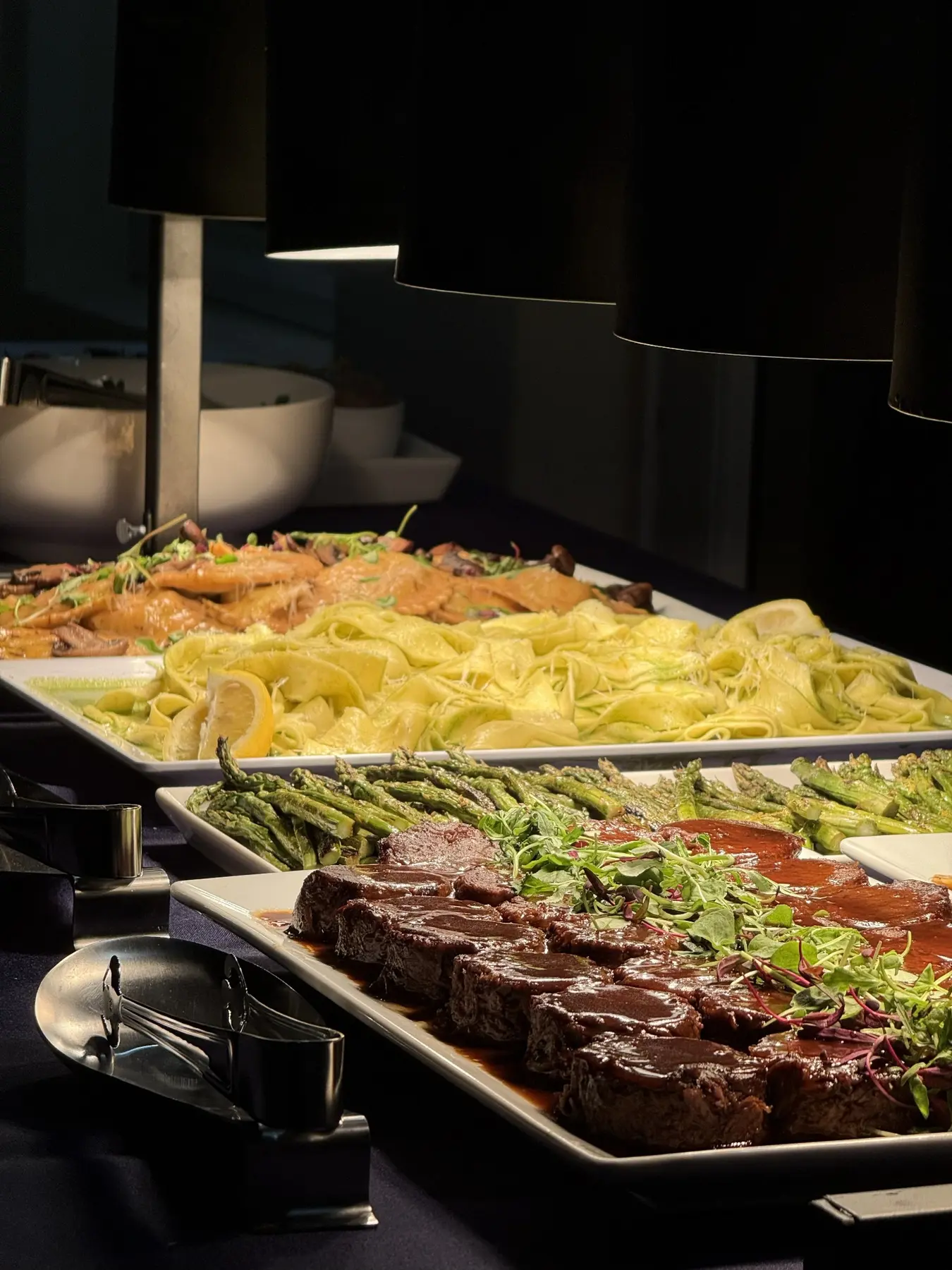 An abundant catered dinner display with beef, pasta and vegetables under warm service lights