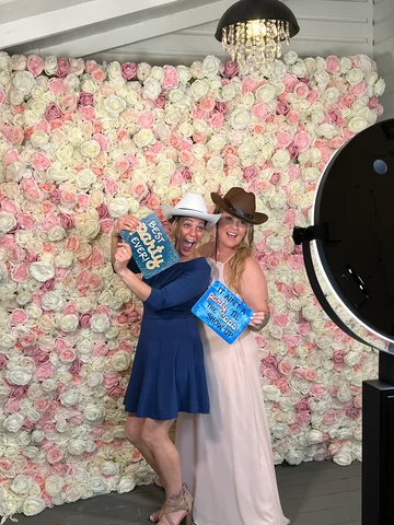 Two girl friends posing for a photo booth in front of a pink and white flower wall wearing cowboy hats and holding signs