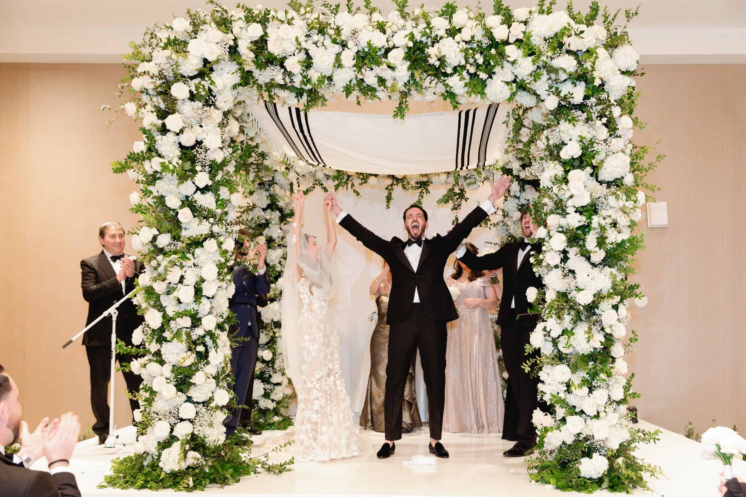 Bride and groom celebrating under a floral arch with friends clapping and cheering.
